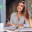 Femme souriante rédigeant des notes sur un carnet, ordinateur et tasse de café à portée de main.