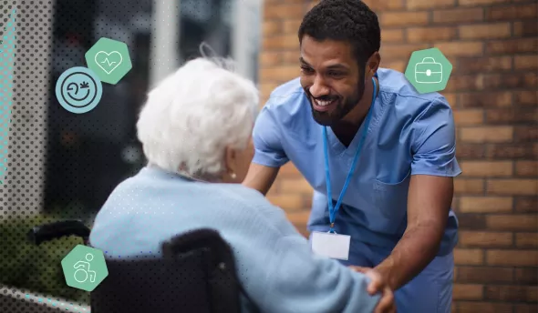 Aidant souriant échange avec une personne âgée en fauteuil roulant devant un bâtiment.